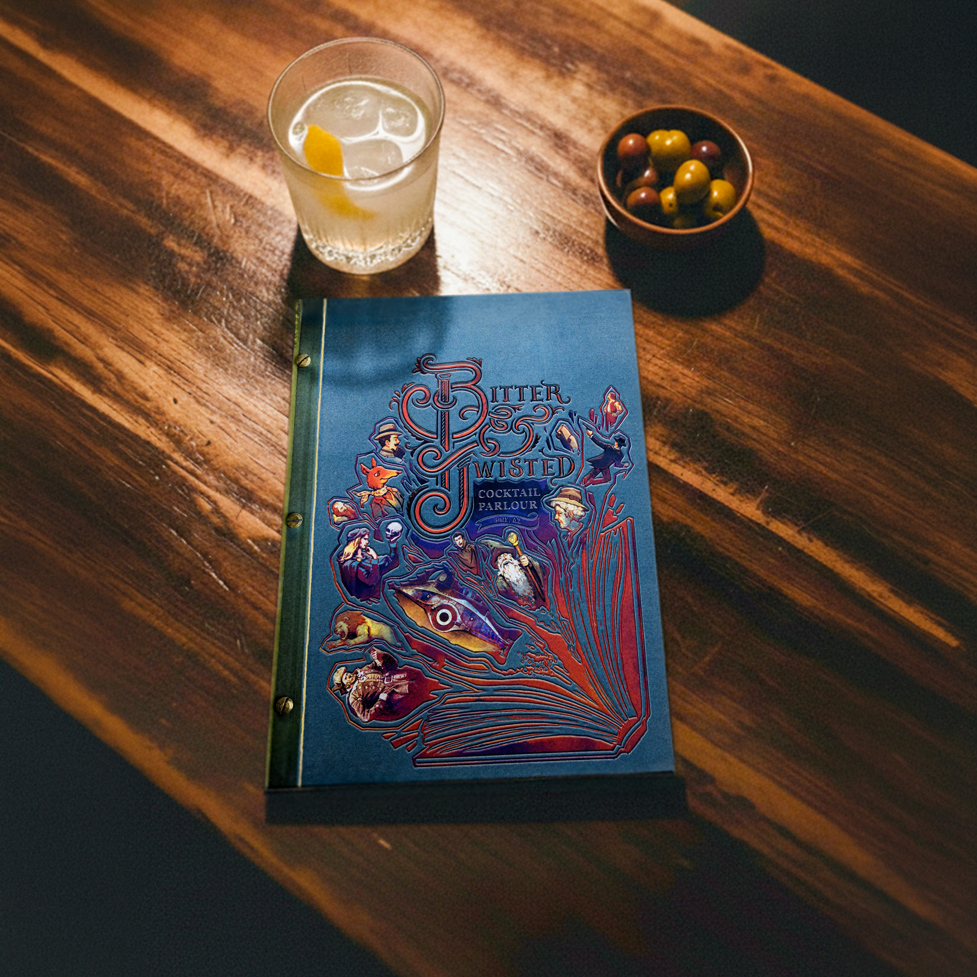 Overhead view of a Bitter and Twisted menu on a wood table, next to a cocktail and small bowl of olives